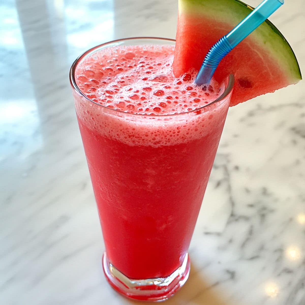 Tall glass of homemade watermelon juice with frothy top, blue straw, and watermelon wedge.