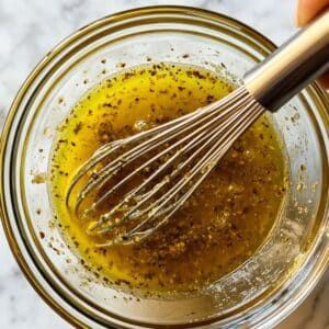 Whisking homemade Greek salad dressing with olive oil and lemon. Bright, herby emulsion for flavorful pasta salads.