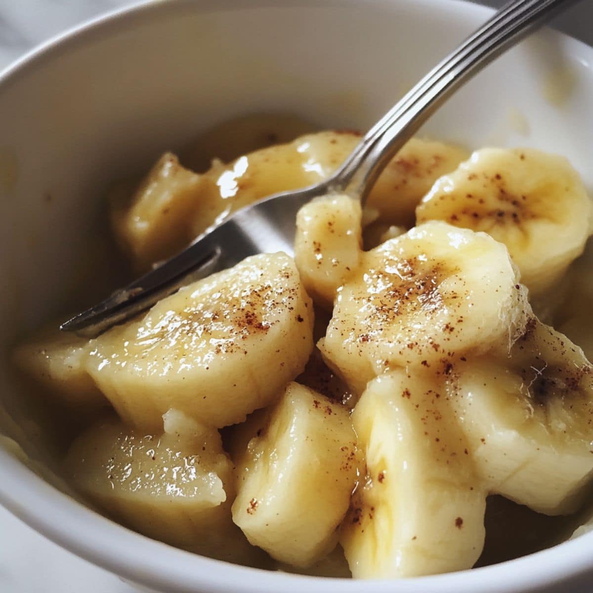 Mashed ripe bananas with brown spots in a white bowl on a white marble counter.