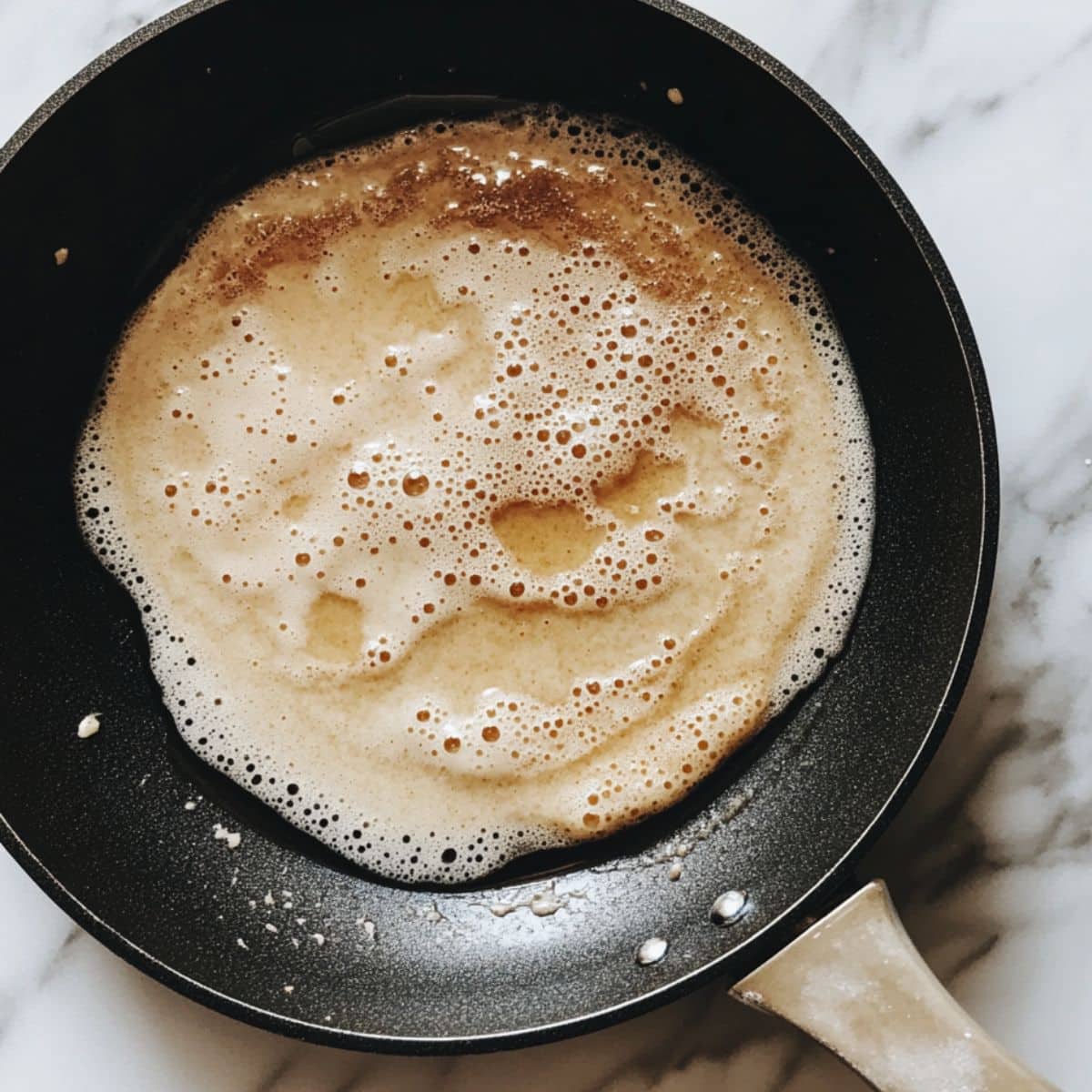 Banana pancake batter cooking on skillet with bubbles forming and golden edges. Homemade breakfast in progress on white stovetop.