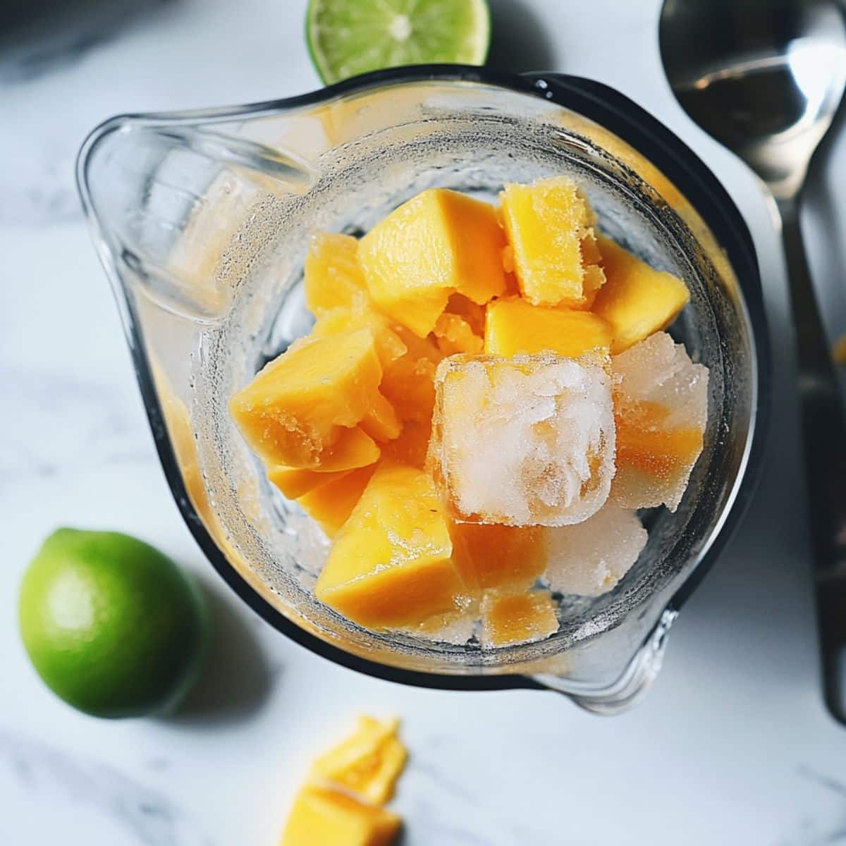 Unblended mango margarita mix in blender with mango, tequila, ice, and lime juice on a white counter.