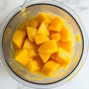 Mango chunks blending into golden puree for sorbet. Natural, homemade process on a white kitchen surface.