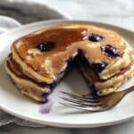 Fluffy blueberry pancakes stacked on a white plate, bursting with juicy berries and golden edges, with a wedge cut out to show the soft center and syrup drizzle.