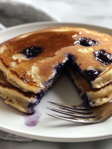 Fluffy blueberry pancakes stacked on a white plate, bursting with juicy berries and golden edges, with a wedge cut out to show the soft center and syrup drizzle.