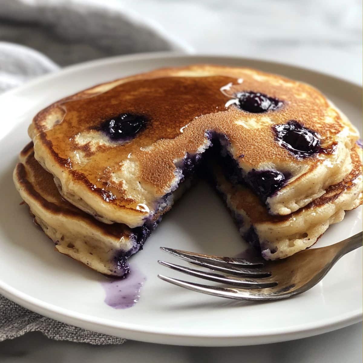 Fluffy blueberry pancakes stacked on a white plate, bursting with juicy berries and golden edges, with a wedge cut out to show the soft center and syrup drizzle.