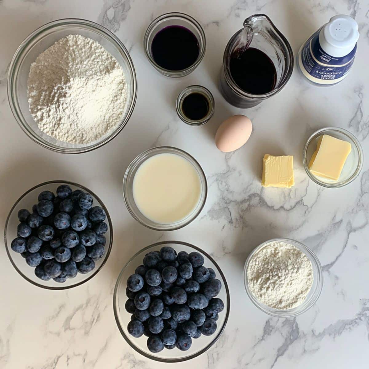 All ingredients for homemade blueberry waffles laid out on a white kitchen counter. Includes flour, eggs, buttermilk, blueberries, and maple syrup.