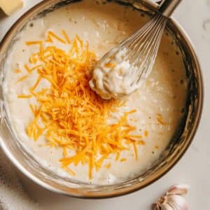Cheddar cheese melting into creamy garlic sauce on stovetop for au gratin potatoes.