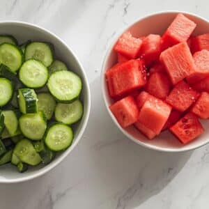 Diced cucumbers salted in one bowl and juicy red watermelon cubes in another. Early prep for a fresh summer salad.