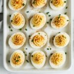 Overhead shot of classic deviled eggs on a white tray, garnished with paprika and chives. Homemade texture with creamy, swirled filling.