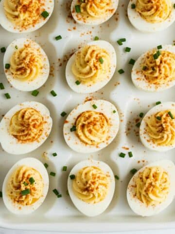 Overhead shot of classic deviled eggs on a white tray, garnished with paprika and chives. Homemade texture with creamy, swirled filling.