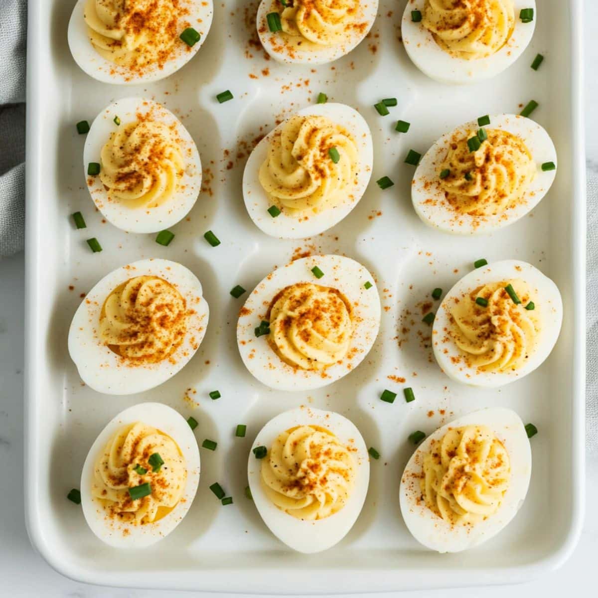 Overhead shot of classic deviled eggs on a white tray, garnished with paprika and chives. Homemade texture with creamy, swirled filling.