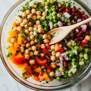 Three bean salad being gently tossed in a glass bowl. Juicy colorful ingredients coated in vinaigrette, final step before chilling.