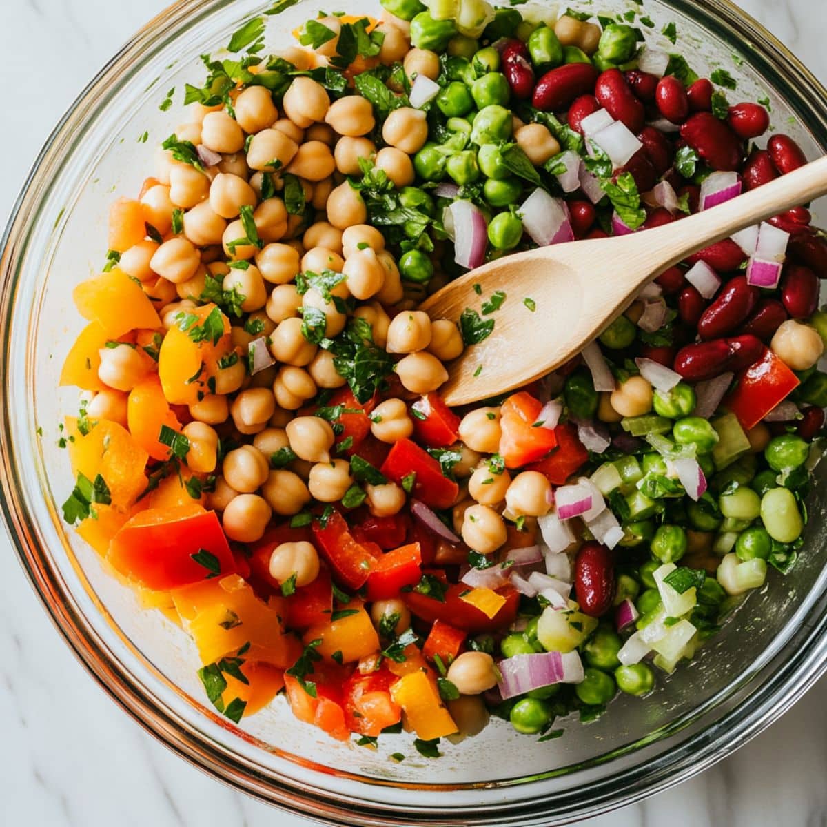 Three bean salad being gently tossed in a glass bowl. Juicy colorful ingredients coated in vinaigrette, final step before chilling.