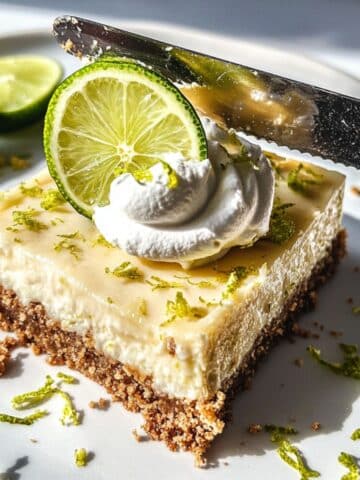 Chilled knife slicing key lime bars on a white kitchen counter. Topped with whipped cream and lime zest.