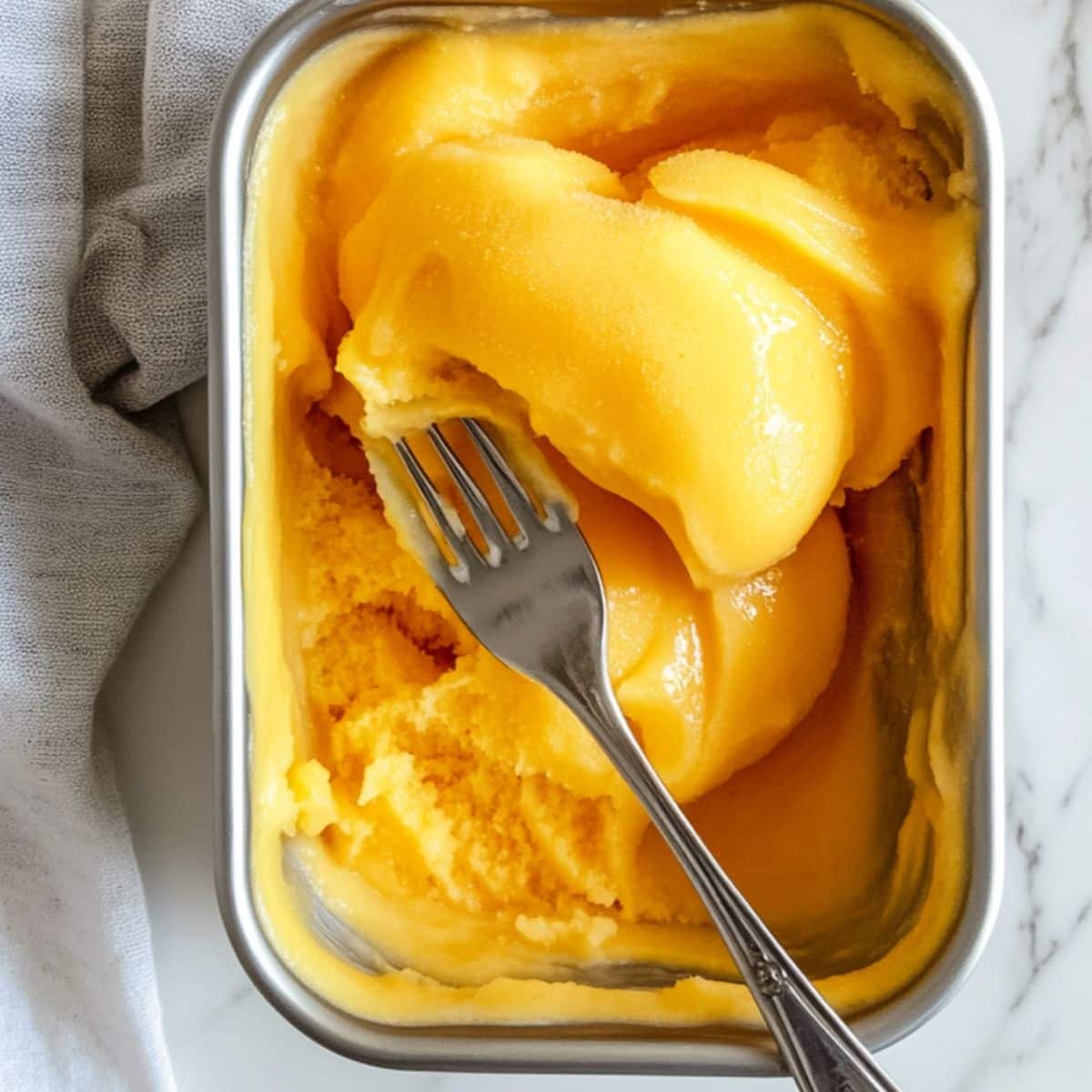 Partially frozen mango sorbet being stirred with fork. Homemade no-churn method on white kitchen counter.