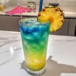 Layered mermaid cocktail with blue, green, and yellow tones, garnished with pineapple and umbrella on a white counter.