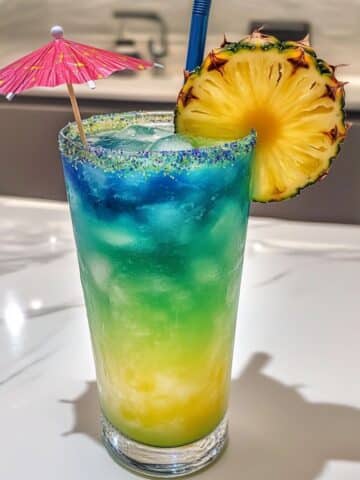 Layered mermaid cocktail with blue, green, and yellow tones, garnished with pineapple and umbrella on a white counter.