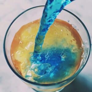 Blue curaçao layer being poured over bar spoon to create gradient in mermaid cocktail. Homemade vibe with visible layering.