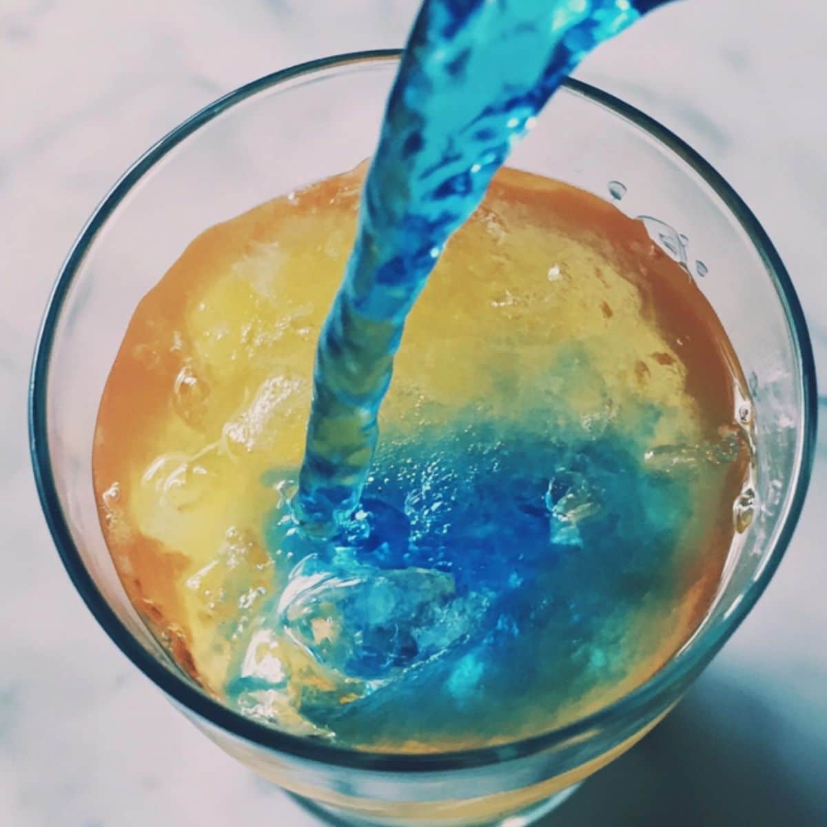 Blue curaçao layer being poured over bar spoon to create gradient in mermaid cocktail. Homemade vibe with visible layering.