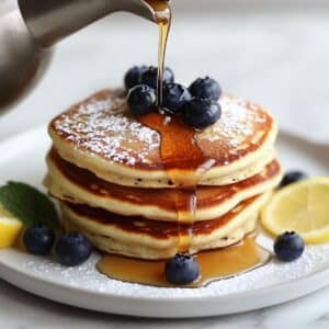 Fluffy ricotta pancakes stacked with blueberries, powdered sugar, and maple syrup. A homemade breakfast served with lemon on a white plate.