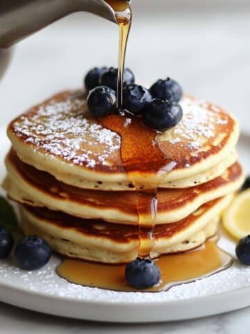 Fluffy ricotta pancakes stacked with blueberries, powdered sugar, and maple syrup. A homemade breakfast served with lemon on a white plate.