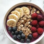 Homemade acai bowl with berries, granola, banana, and almond butter on a white kitchen counter. Realistic and imperfectly styled.