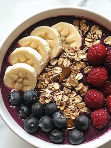 Homemade acai bowl with berries, granola, banana, and almond butter on a white kitchen counter. Realistic and imperfectly styled.