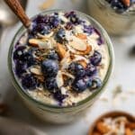 Close-up of homemade blueberry overnight oats in a jar with coconut and chopped almonds. Creamy and vibrant, served on white marble countertop.