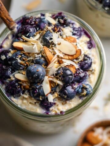 Close-up of homemade blueberry overnight oats in a jar with coconut and chopped almonds. Creamy and vibrant, served on white marble countertop.