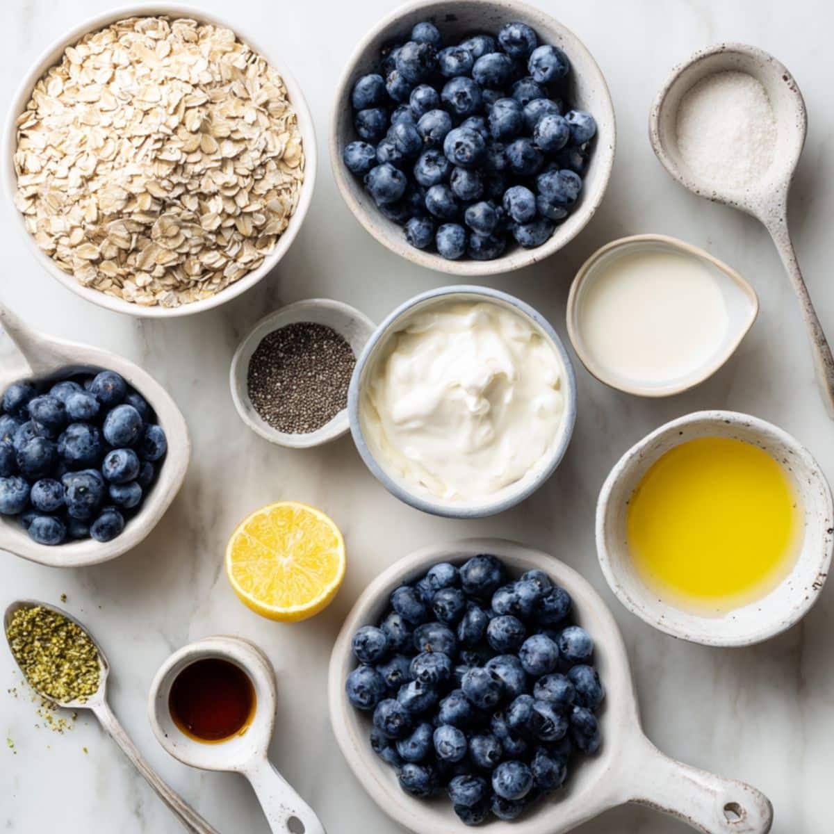 All ingredients for blueberry overnight oats laid out casually on white counter. Includes oats, yogurt, blueberries, chia, maple, lemon zest.