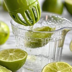 Juicing fresh limes by hand for cherry limeade. Lime juice collecting in a glass cup on white counter.