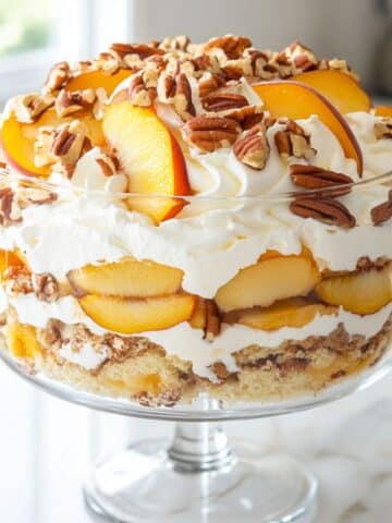 Peach trifle topped with thick whipped cream and toasted pecans. Luscious layers of peaches, cake, and cream inside a clear bowl.