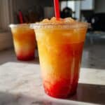 Refreshing pineapple passionfruit lemonade blended with strawberry purée in a clear cup, set on a white marble kitchen counter with vibrant layered colors and icy texture.