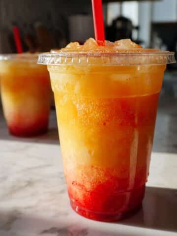 Refreshing pineapple passionfruit lemonade blended with strawberry purée in a clear cup, set on a white marble kitchen counter with vibrant layered colors and icy texture.