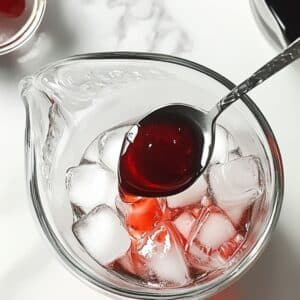 Cherry syrup being poured into a pitcher with ice for limeade. Homemade step on white kitchen counter.