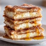 Stacked sopapilla cheesecake bars with flaky pastry and creamy filling. Cinnamon sugar top and honey drizzle on white plate.
