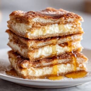 Stacked sopapilla cheesecake bars with flaky pastry and creamy filling. Cinnamon sugar top and honey drizzle on white plate.