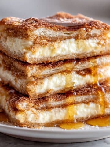 Stacked sopapilla cheesecake bars with flaky pastry and creamy filling. Cinnamon sugar top and honey drizzle on white plate.