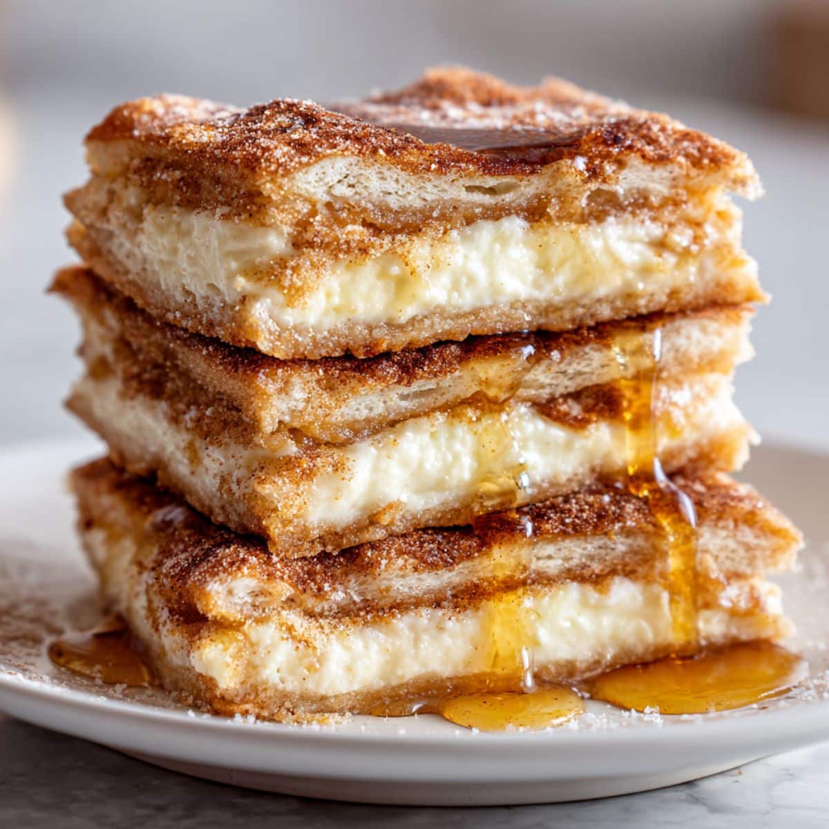 Stacked sopapilla cheesecake bars with flaky pastry and creamy filling. Cinnamon sugar top and honey drizzle on white plate.