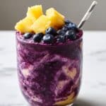 Vibrant blueberry and pineapple smoothie swirled in a clear glass, topped with fresh pineapple chunks and blueberries, set on a white marble kitchen counter for a refreshing, juicy close-up.
