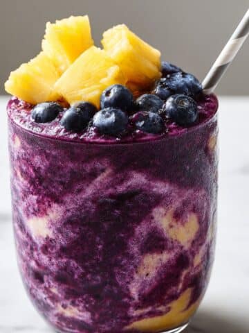 Vibrant blueberry and pineapple smoothie swirled in a clear glass, topped with fresh pineapple chunks and blueberries, set on a white marble kitchen counter for a refreshing, juicy close-up.