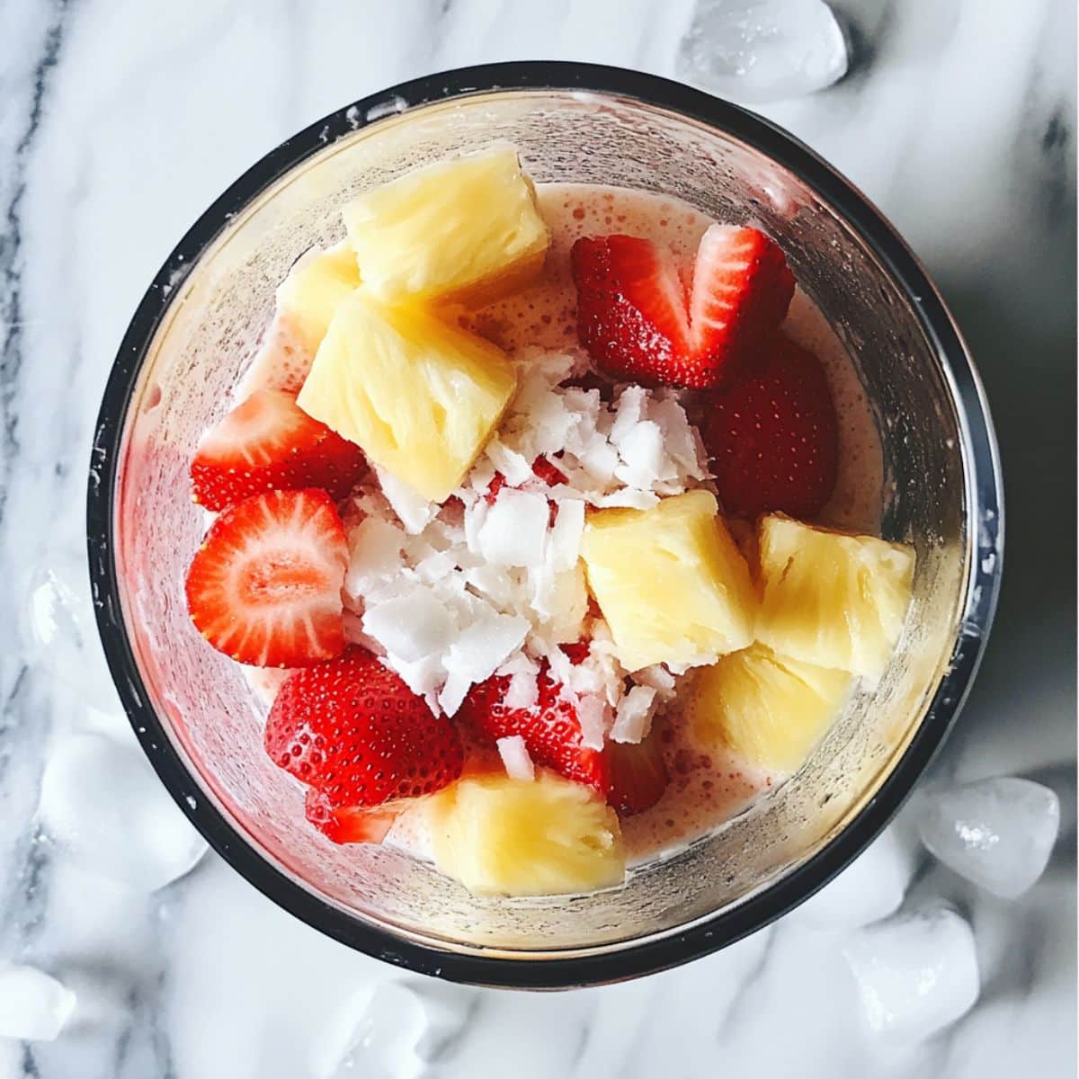 Strawberries, pineapple, and coconut cream being blended into pink Strawberry piña colada. Homemade drink prep on white counter.