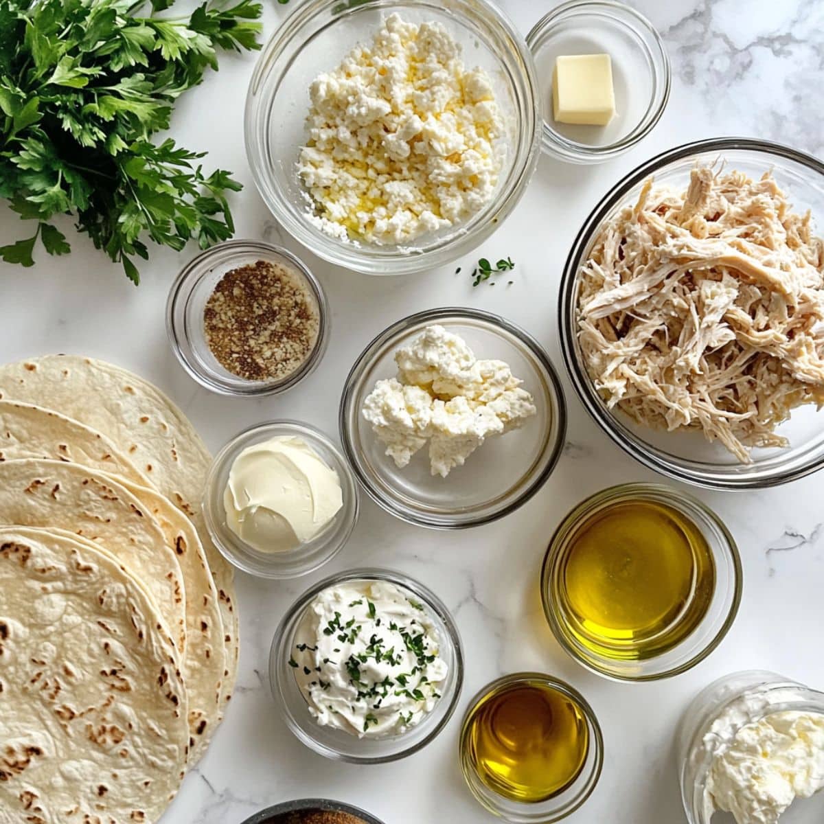 All ingredients for cheesy garlic chicken wraps displayed in bowls on a white counter. Includes chicken, cheeses, garlic, tortillas, and seasonings.