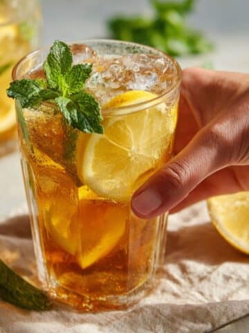 Hand holding a homemade glass of mango iced tea with ice, mint, and lemon.