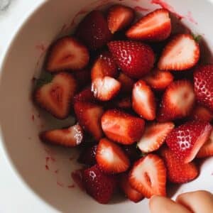 Fresh strawberries cut into quarters with berry juice on counter. A child's hand helps prep on a real home kitchen surface.