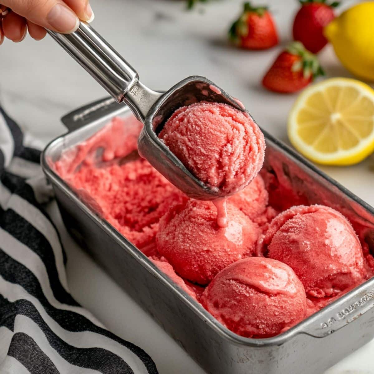 A hand scooping homemade strawberry sorbet from a loaf pan. Natural texture and rustic kitchen setup on wooden counter with striped towel.