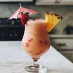 Naturally pink strawberry piña colada served in a glass with fruit garnish. Homemade tropical drink on white kitchen counter.
