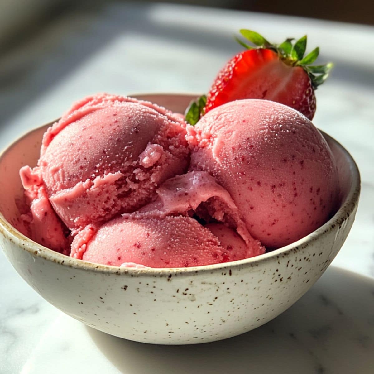 Three scoops of homemade strawberry sorbet in a rustic white bowl with a fresh strawberry slice. Naturally styled on a white kitchen counter.