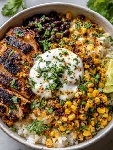 Homemade street corn chicken rice bowl with juicy grilled chicken, corn, beans, rice, and crema in white bowl.