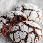 Homemade chocolate crinkle cookies with white powdered sugar cracks, one bitten showing fudgy interior Three chocolate crinkle cookies on parchment paper displaying perfect cracking and brownie-like texture